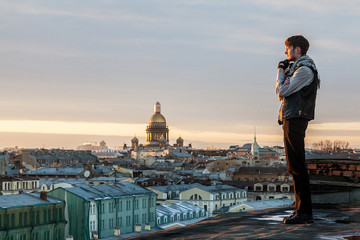 A bearded guy in a leather vest and jeans stands on the edge of the roof with a view of St. Petersburg at sunset