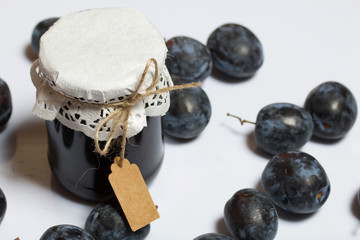 Jar of plum jam. With an inscription tag. Plums are scattered nearby. On a white background.