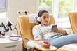 © AnnaStills - Young woman in headphones using her mobile phone to listen to music while lying on the couch and donating blood