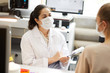 © AnnaStills - Female doctor in mask making notes in medical card while talking to patient at hospital