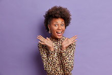 Happy Dark Skinned Woman With Curly Natural Hair, Raises Palms From Joy, Smiles Broadly, Hears Excellent News, Wears Leopard Jumper, Models Against Vivid Purple Background, Amused By Someone