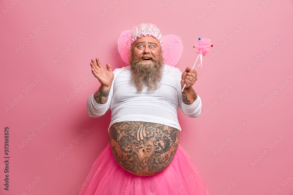 Horizontal shot of joyous funny man with thick beard and big fat belly, plays fairy on costume party, chills during spare time, sends positive vibes, works as animator, isolated over rosy background - obrazy, fototapety, plakaty 