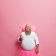 © wayhome.studio  - Funny plump daddy plays fairy on birthday party, amuses children, foolishes around, looks hopefully above as makes wish with magic wand, pink background. Man acts out fairytale has fun plays with kids