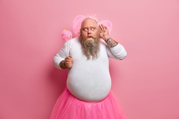 Funny fatso man in princess outfit, holds magic wand, wears pink wings, has big fat belly, keeps hand near ear, tries to hear something interesting, has curious expression, isolated on rosy background