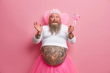 horizontal shot of joyous funny man with thick beard and big fat belly, plays fairy on costume party