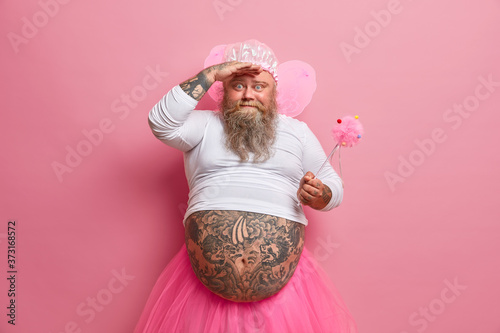 Photo of funny bearded father dressed in carnival costume, plays with children, wears fairy wings, undersized jumper and rosy pleated skirt, holds magic wand Tableau sur Toile
