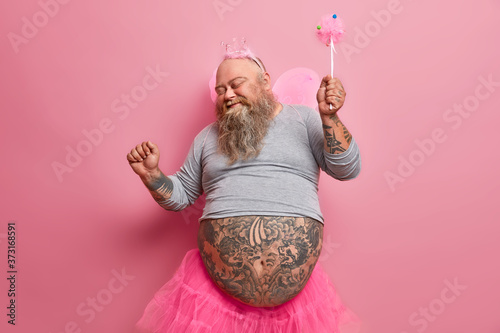 Fotografía Overjoyed stout man in fairy costume dances carefree, has fun and foolishes around, organises holiday for children, plays princess, moves against pink background
