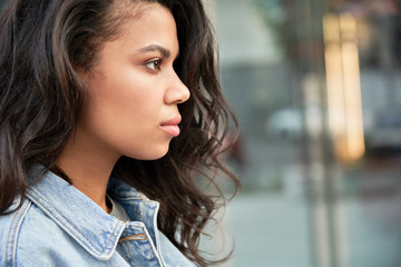 Confident young African American woman looking forward on urban city street, side profile face view. Serious beautiful curly young hipster mixed race teen girl standing posing for headshot portrait.