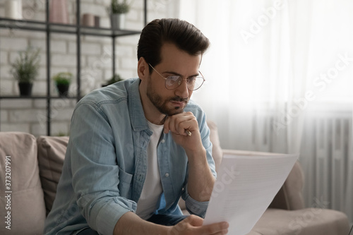 Thoughtful young caucasian man in glasses sit on sofa in living room read postal letter correspondence, serious pensive male analyze consider post paperwork or bank notice, manage budget at home