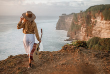Woman Traveler Hold The Guitar Standing On The Cliff And Looking For View Of The Sea And Sunset On Vacation.Travel Concept. Back View