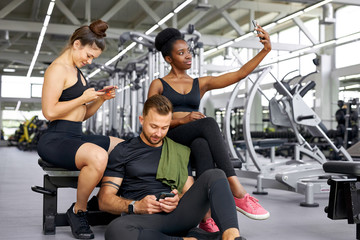 Wall Mural - portrait of tired diverse fit people enjoying time after sport exercises, african and caucasian team sit together in gym, using smartphone