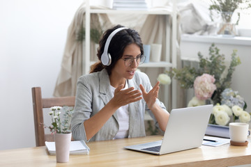 Head shot smiling young creative businesswoman in glasses wearing wireless headphones, enjoying video call conversation with partners or giving professional consultation to client online in showroom.