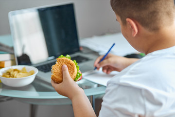 fat overweight boy eat junk food while doing homework, young caucasian boy sit at desk with notebooks, laptop and books. at home