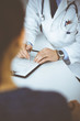 © rogerphoto - A doctor is talking to his patient, while sitting together at the desk in the cabinet in a hospital. Physician using clipboard for filling up medication history records. Perfect medical service in