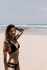 Fit girl in black bikini stands on the beach and holds her hair with her hand.