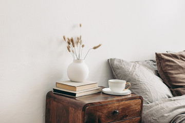 Modern white ceramic vase with dry Lagurus ovatus grass and cup of coffee on retro wooden bedside table. Beige linen and velvet pillows in bedroom. Scandinavian interior. Homestaging.