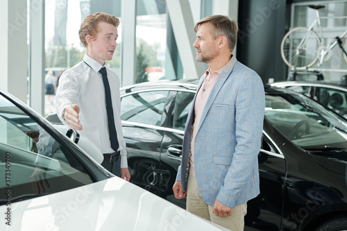 Mature man looking at young sales manager pointing at car and listening to him