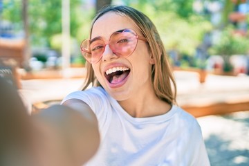 Young beautiful blonde caucasian woman smiling happy outdoors on a sunny day wearing heart shaped sunglasses taking a selfie picture