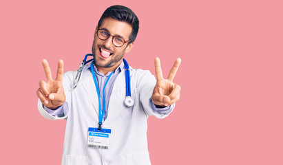 Young hispanic man wearing doctor uniform and stethoscope smiling with tongue out showing fingers of both hands doing victory sign. number two.