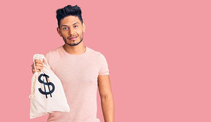 Handsome latin american young man holding money bag with dollar symbol looking positive and happy standing and smiling with a confident smile showing teeth