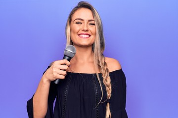 Young beautiful blonde singer woman singing using microphone over purple background looking positive and happy standing and smiling with a confident smile showing teeth