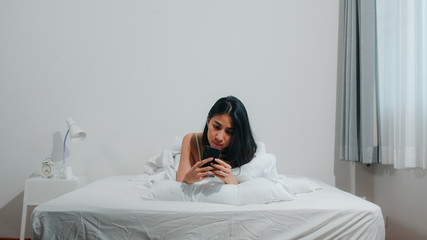 Wall Mural - Young Asian woman using smartphone checking social media feeling happy smiling while lying on bed after wake up in the morning, Beautiful attractive hispanic lady smiling relax in bedroom at home.