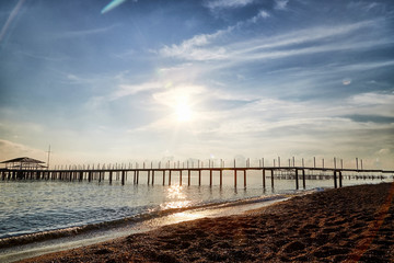 View from sand beach to water of sea, waves and pier in a nice day or evening with blues sky, bright sun and white clouds. The concept of a holiday on the sea or ocean in the South.