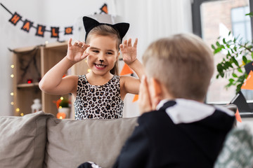 Poster - halloween, holiday and childhood concept - smiling little boy and girl in party costumes playing and having fun at home