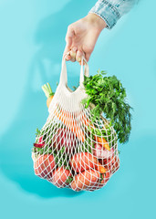 Mesh bag with vegetables and herbs in female hand. Woman hold string net shopping bag on blue background. Modern reusable shopping, eco friendly lifestyle, zero waste concept