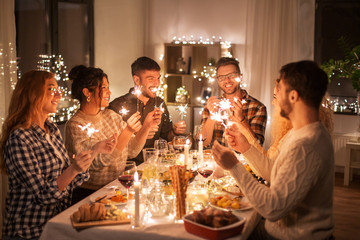 holidays, celebration and people concept - happy friends with sparklers having christmas dinner at h