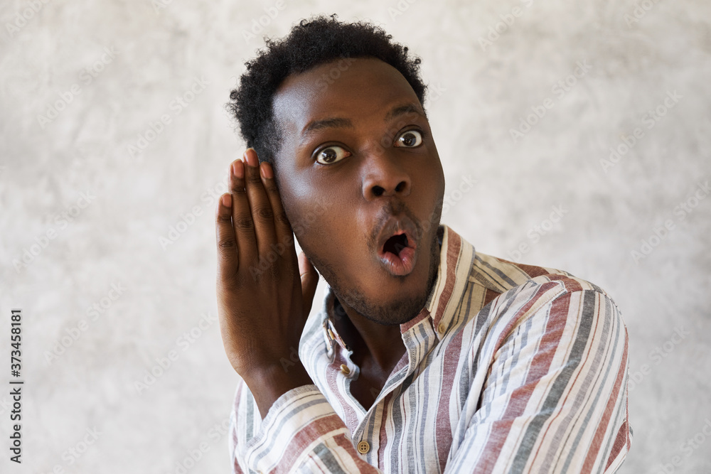 Emotional curious young Afro American man with mouth gaped open expressing surprise and full disbelief holding hand at his ear overhearing secret private conversation, being totally shocked - obrazy, fototapety, plakaty 
