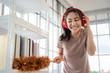 Young asian woman with headphones using a feather duster to clean wood book shelves in home .
