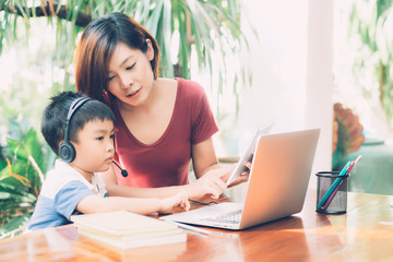 Wall Mural - Young asian mother and son using laptop computer and tablet for study and learn together at home, boy wearing headphone for e-learning with distancing, teacher or mom support child, education concept.