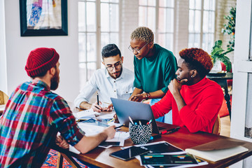 Wall Mural - Group of young hipster IT professionals discussing report information during co brainstorming in modern apartment with wifi connection for developing, diverse students preparing to program test