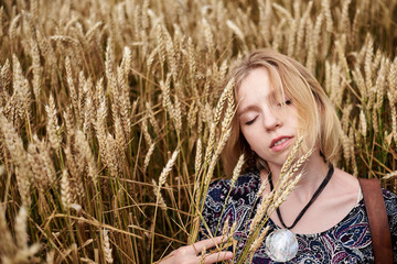 Young blond woman, wearing boho hippie clothes, lying, resting on wheat stalks in the middle of field. Close-up picture of beautiful female face on natural background. Eco tourism. Creative portrait.