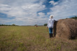 © Юлия Падина - A female farmer stands by a haystack after harvesting in a field with the sky and forest in the background. Landscape