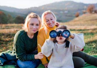 Small girl with mother and grandmother on a walk in autumn nature, using binoculars.