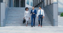 Cheerful Multi Ethnic Young Men And Woman Leaving Business Office Building And Stepping Down The Steps. Outdoors. African American Businesswoman And Mixed-races Businessmen Talking And Using Tablet.