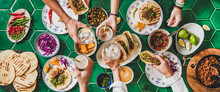Friends Home Taco Party. Flat-lay Of Mexican Traditional Dishes Tacos With Beef Meat, Corn Tortillas , Tomato Salsa And Peoples Hands With Glasses Over Green Background, Top View. Mexican Cuisine