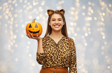 Wall Mural - holiday, theme party and people concept - happy smiling woman in halloween costume of leopard with ears holding jack-o-lantern pumpkin over festive lights background