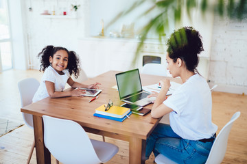 Black mother working on laptop and smiling daughter playing on tablet while looking at camera