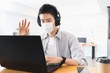 © Ann Patchanan - Young smart asian business man in casual shirt at the office wearing medical face mask under the outbreak of the virus,Greeting colleagues through video conferencing.