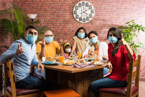 Indian family wears face mask while eating food in restaurant after corona pandemic unlock - concept showing new normal lifestyle in India
