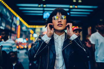 Wall Mural - Half length portrait of trendy female millennial in electronic spectacles looking at camera during time for listening audio book and walk in metropolitan downtown, generation z in earphones