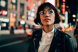 Caucasian hipster girl in classic spectacles for provide eyes protection thinking about metropolitan nightlife in New York, thoughtful female in fashionable outfit recreating during evening in city