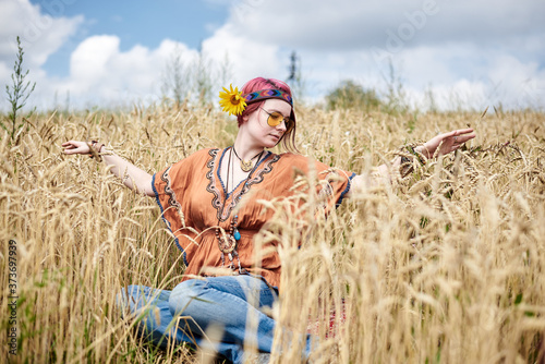 Young hippie woman with red burgundy hair, wearing boho style clothes, sitting in middle of wheat field, meditating, thinking, relaxing, enjoying. Eco tourism concept. Nature protection.