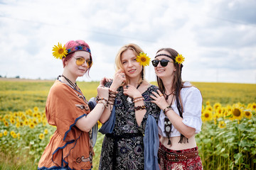 Three young women, wearing boho hippie clothes, standing in the middle of yellow sunflowers field. Girlfriends, traveling together in countryside. Eco tourism concept.