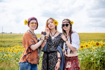 Three young women, wearing boho hippie clothes, standing in the middle of yellow sunflowers field. Girlfriends, traveling together in countryside. Eco tourism concept.