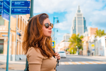 A young woman in sunglasses stands at a traffic light on a business street in Moscow on a summer day