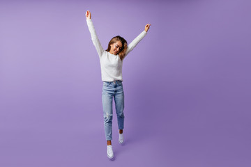 Wall Mural - Cheerful student in good mood happily jumping in studio on isolated background. Long-haired brown-haired woman in stylish jeans and white sneakers poses for full-length photo